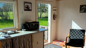 A compact kitchen with a refrigerator, a microwave, and a kettle on the counter. There is a dining table with chairs, a small window, and a door leading outside. - Charming summer house in an idyllic area. (Odder Gylling)