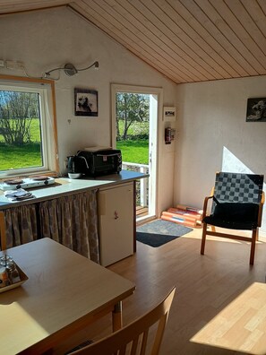A compact kitchen with a refrigerator, a microwave, and a kettle on the counter. There is a dining table with chairs, a small window, and a door leading outside.