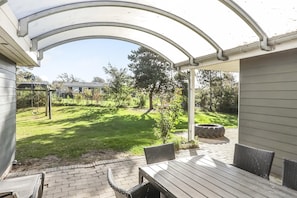 Un patio cubierto con mesa y sillas, un camino de ladrillo y vistas a un jardín con césped, árboles y un columpio.