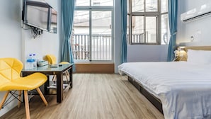 Habitación de hotel con una cama, un escritorio y sillas, un televisor montado en la pared y una ventana con cortinas.