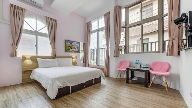 Un dormitorio con cama, escritorio, dos sillas y ventanas amplias con cortinas.