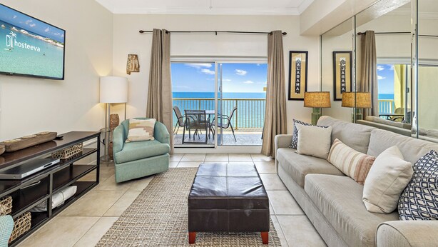A spacious living room with a large window offering a view of the ocean, a ceiling fan, and a flat-screen TV mounted on the wall. - Royal Palms Condos (Gulf Shores)