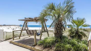 A wooden swing set with a bench, surrounded by palm trees and a white fence, overlooking a beach.