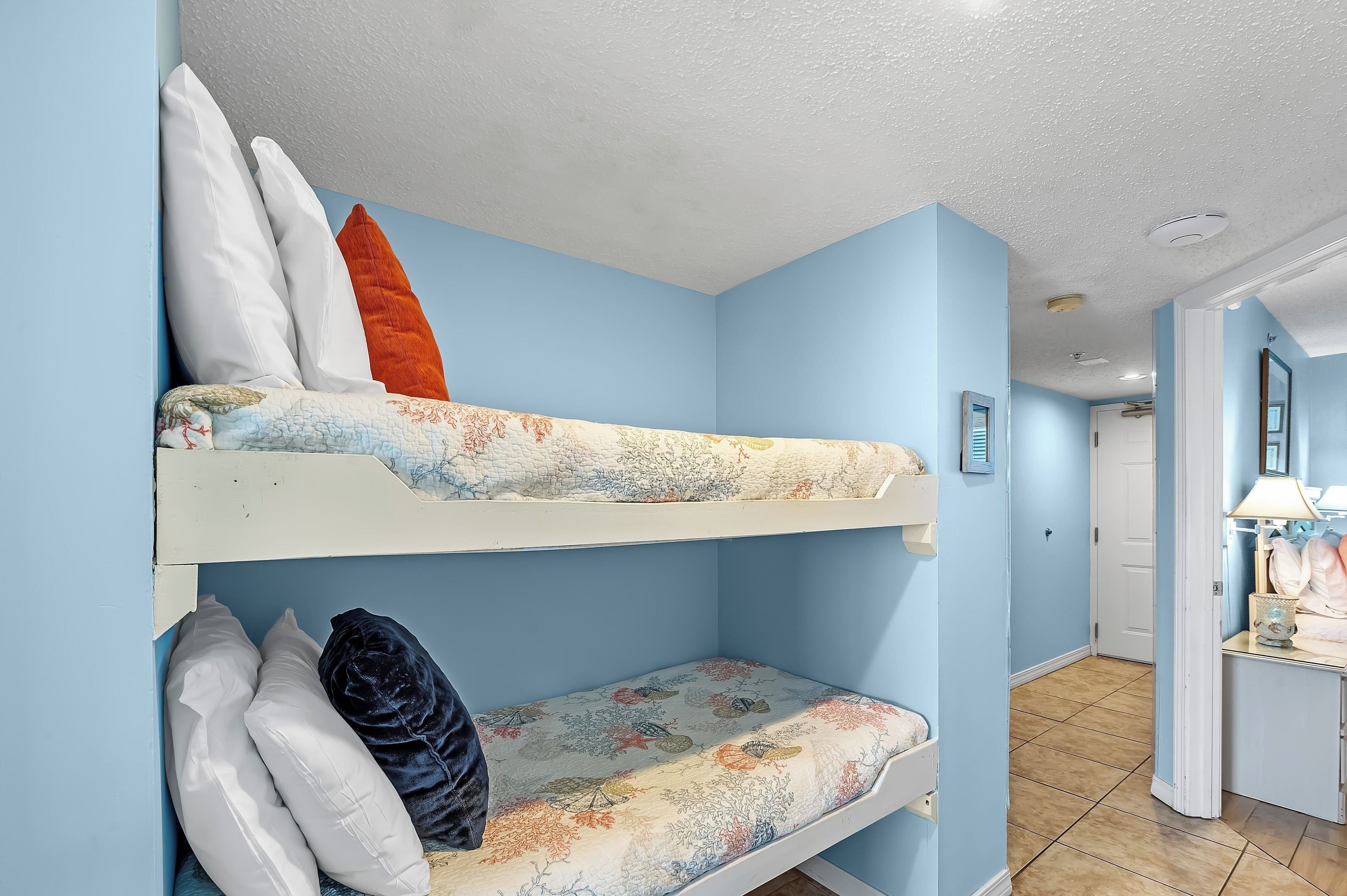 Een stapelbed met bloemenbeddengoed en kussens, een klein nachtkastje met een lamp, en een deur naar een andere kamer.