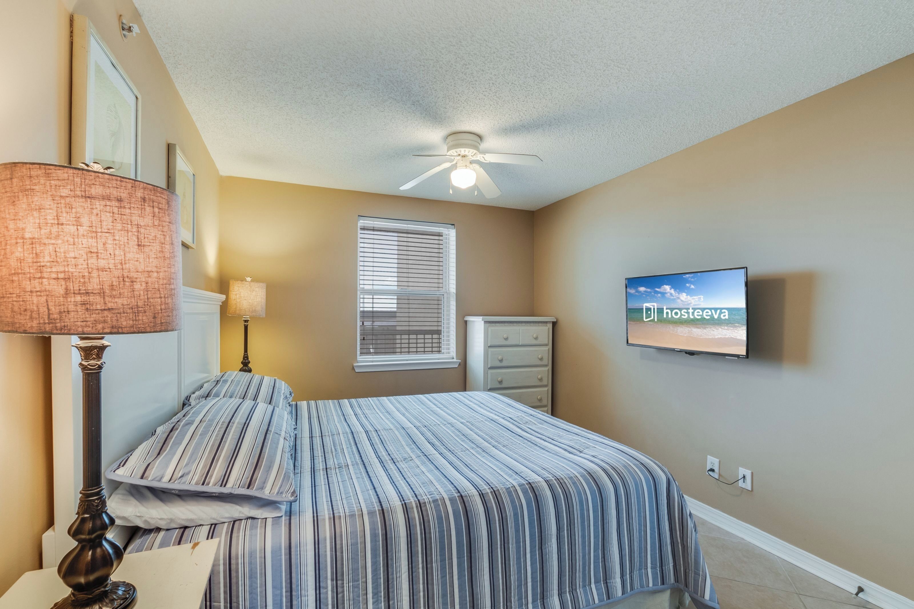 A bedroom with a bed, a ceiling fan, a TV, a window with blinds, and a lamp.