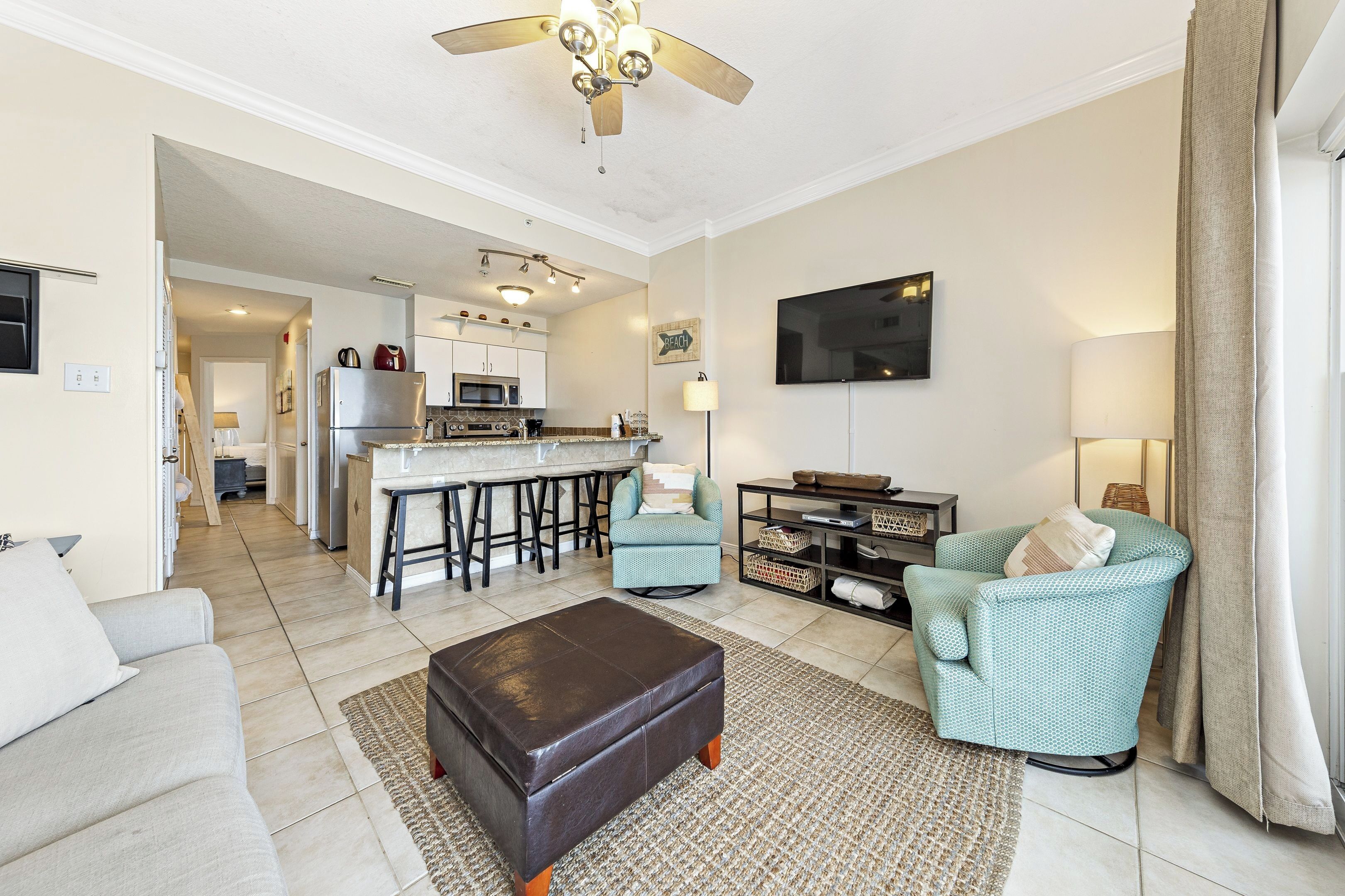 A modern living room with a sofa, ottoman, and teal armchairs, a kitchen area with a bar, and a ceiling fan.