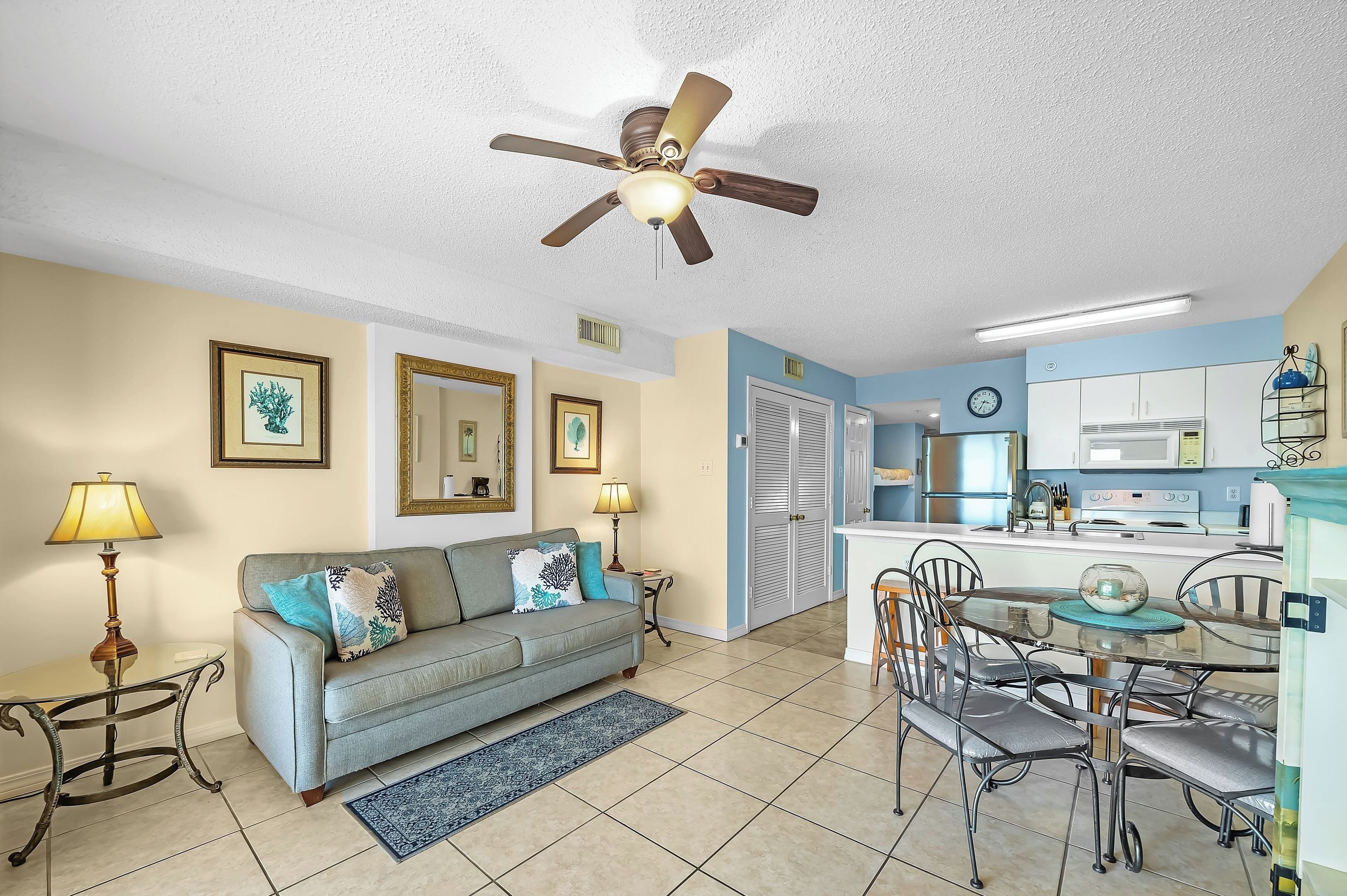 A living room with a sofa, a dining table with chairs, a ceiling fan, and a kitchen area with cabinets and appliances.