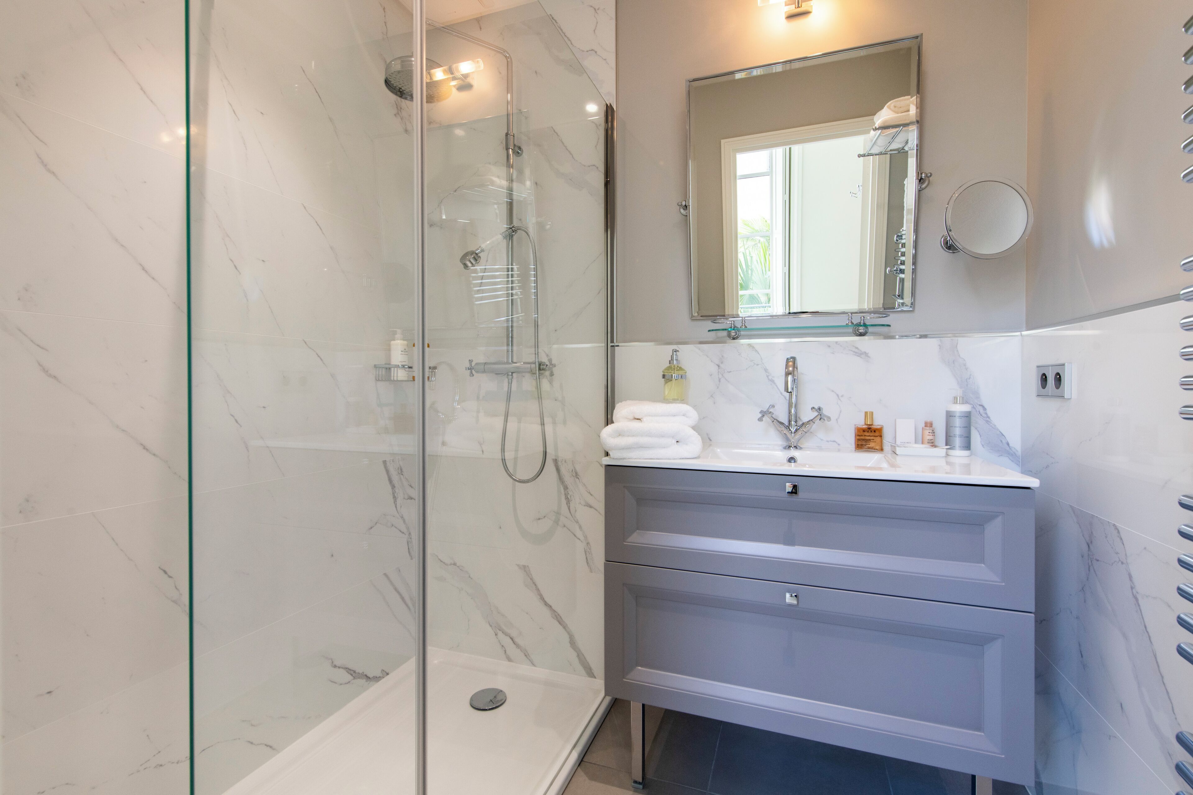 A modern bathroom with a glass-enclosed shower, a marble-tiled wall, a sink with a mirror, and a vanity with drawers.