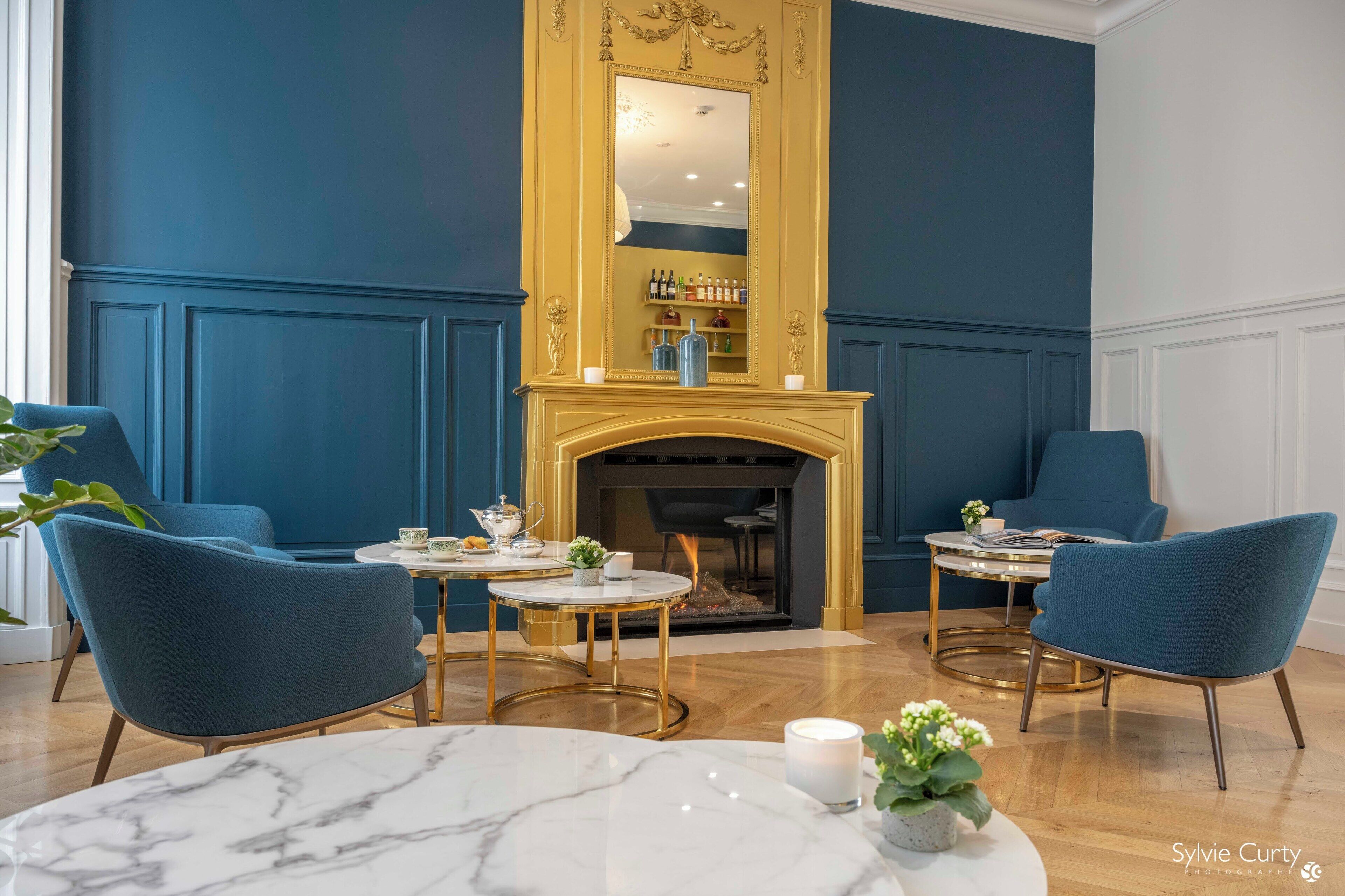 Una habitación con chimenea, dos sillones azules, una mesa de mármol y un espejo con marco dorado.