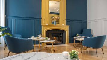 Una habitación con chimenea, dos sillones azules, una mesa de mármol y un espejo con marco dorado.