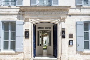 Una entrada imponente con una puerta blanca y contraventanas azules, que conduce a un patio con una fuente y vegetación.