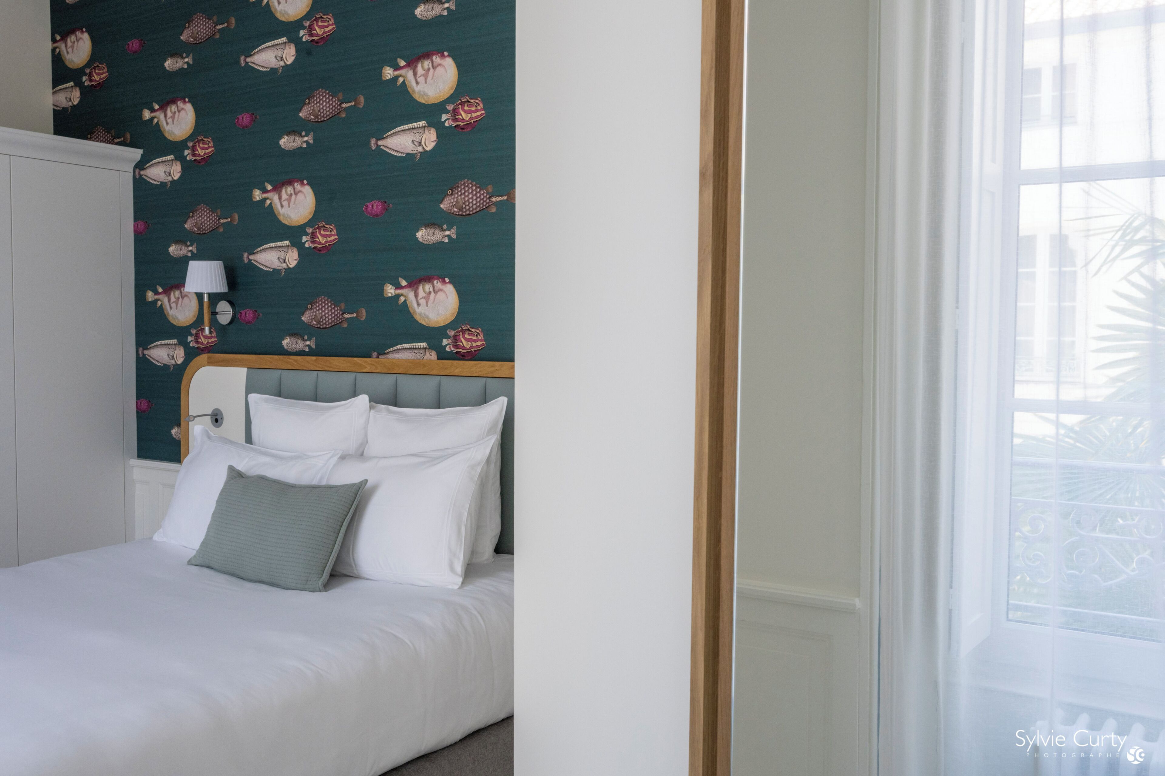 A neatly made bed with white and green pillows, a teal patterned wall, and a window with a view of buildings.