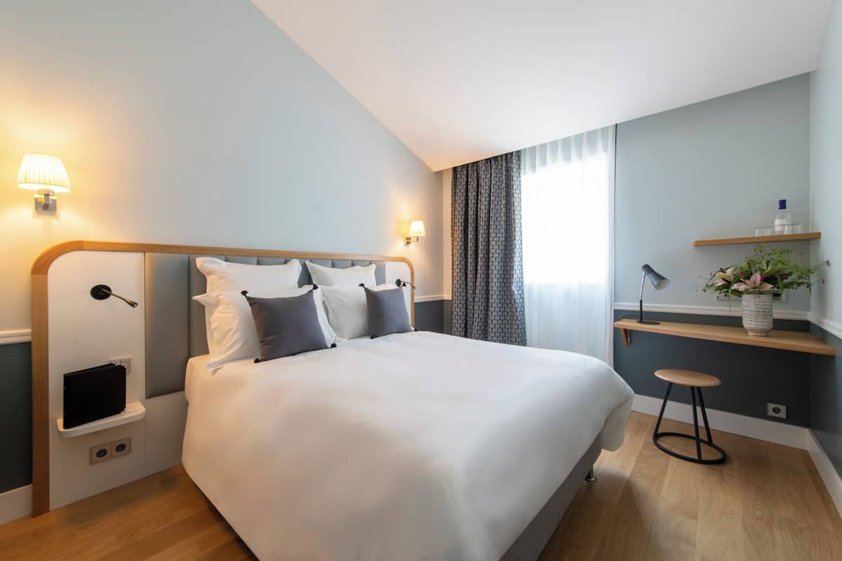 A modern hotel room with a large bed, bedside tables, a small stool, a nightstand, a wall-mounted lamp, and a shelf with a vase of flowers.