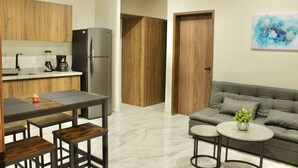 A modern kitchen with wooden cabinets, a refrigerator, and a coffee maker. There is a dining table with benches and a gray sofa with a plant.