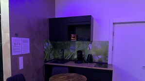 Eine kompakte Küchenzeile mit Kaffeemaschine, kleinem Kühlschrank und Spüle. Ein einzelner Stuhl und ein kleiner runder Tisch gehören ebenfalls dazu.