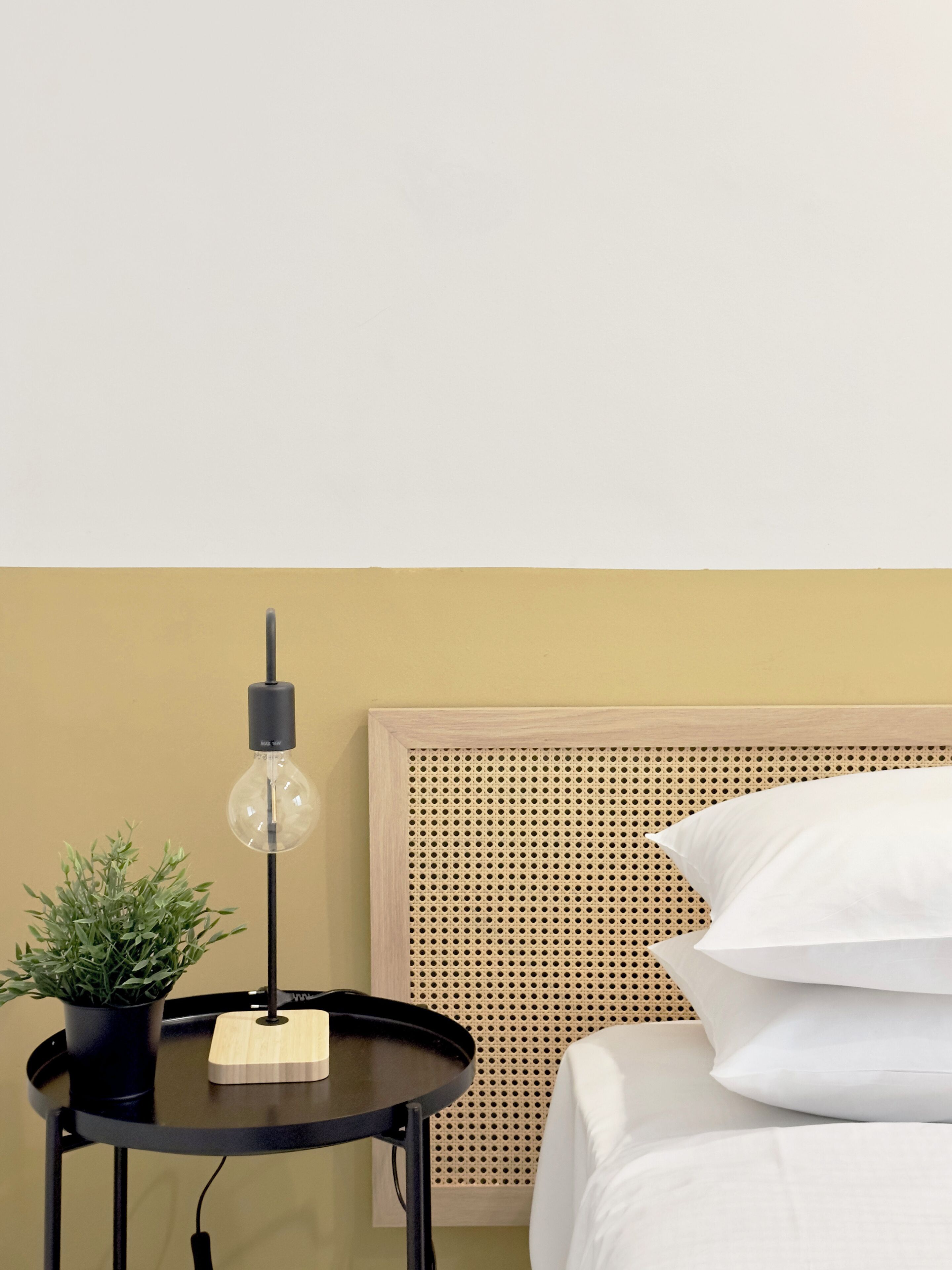 A neatly made bed with white pillows and a headboard with a perforated design. A small round table with a black lamp and a potted plant is beside the bed.