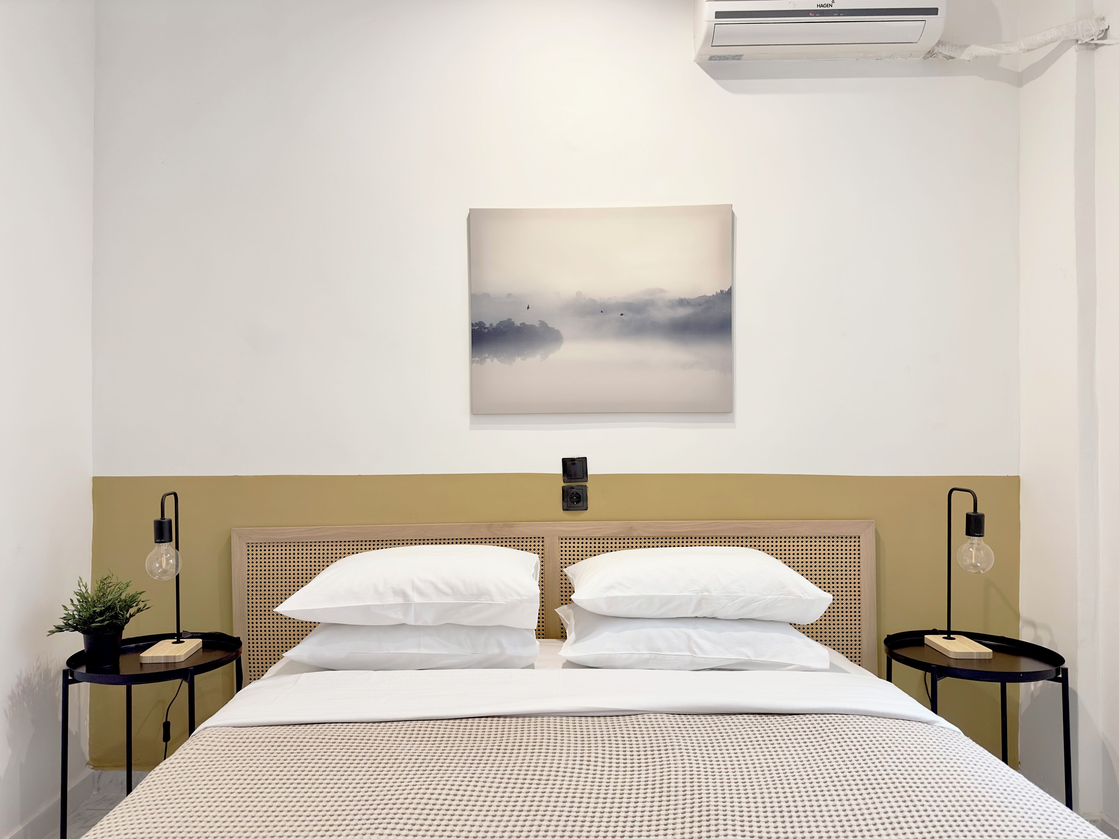 A neatly made bed with a headboard, two bedside tables with lamps, a wall-mounted air conditioner, and a framed picture above the bed.