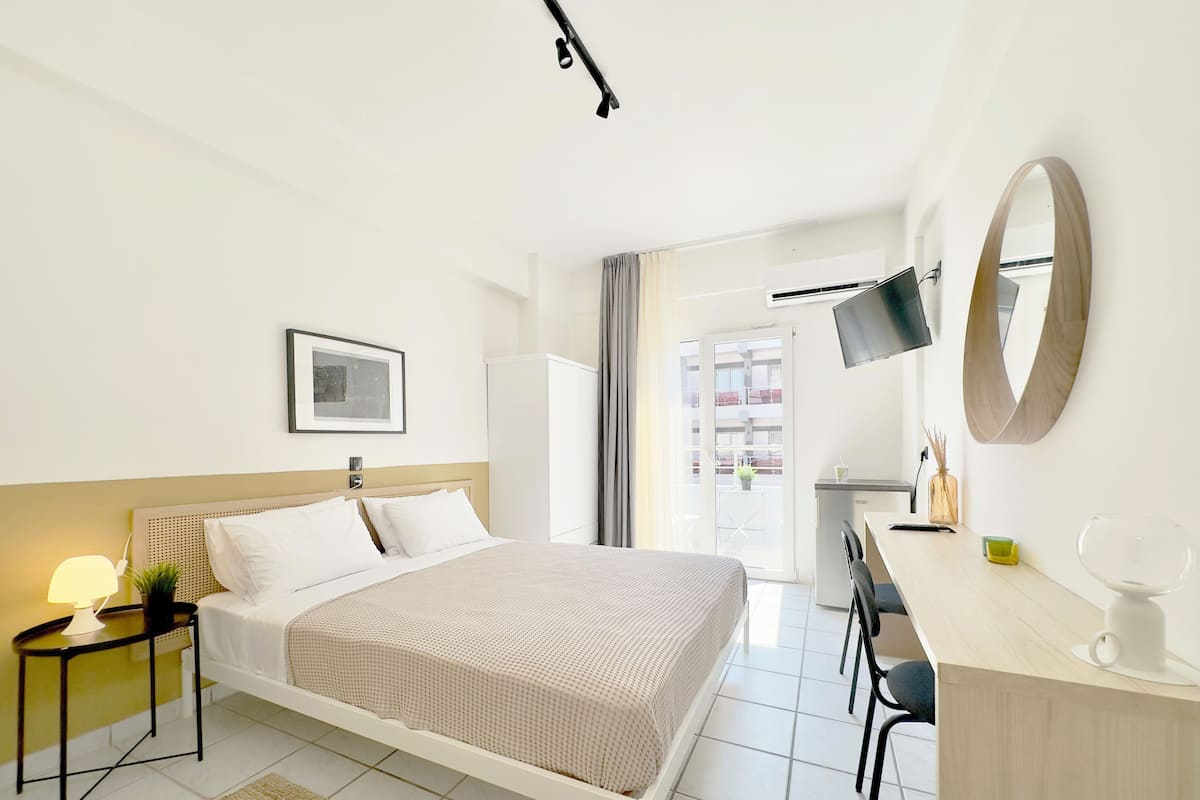 A hotel room with a bed, bedside table, lamp, desk with chair, TV, mirror, and a view of the outside balcony.