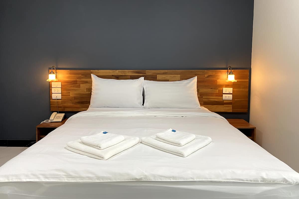Un lit bien fait, avec du linge de lit blanc et deux serviettes pliées, une tête de lit en bois, des lampes de chevet et un téléphone sur la table de chevet.
