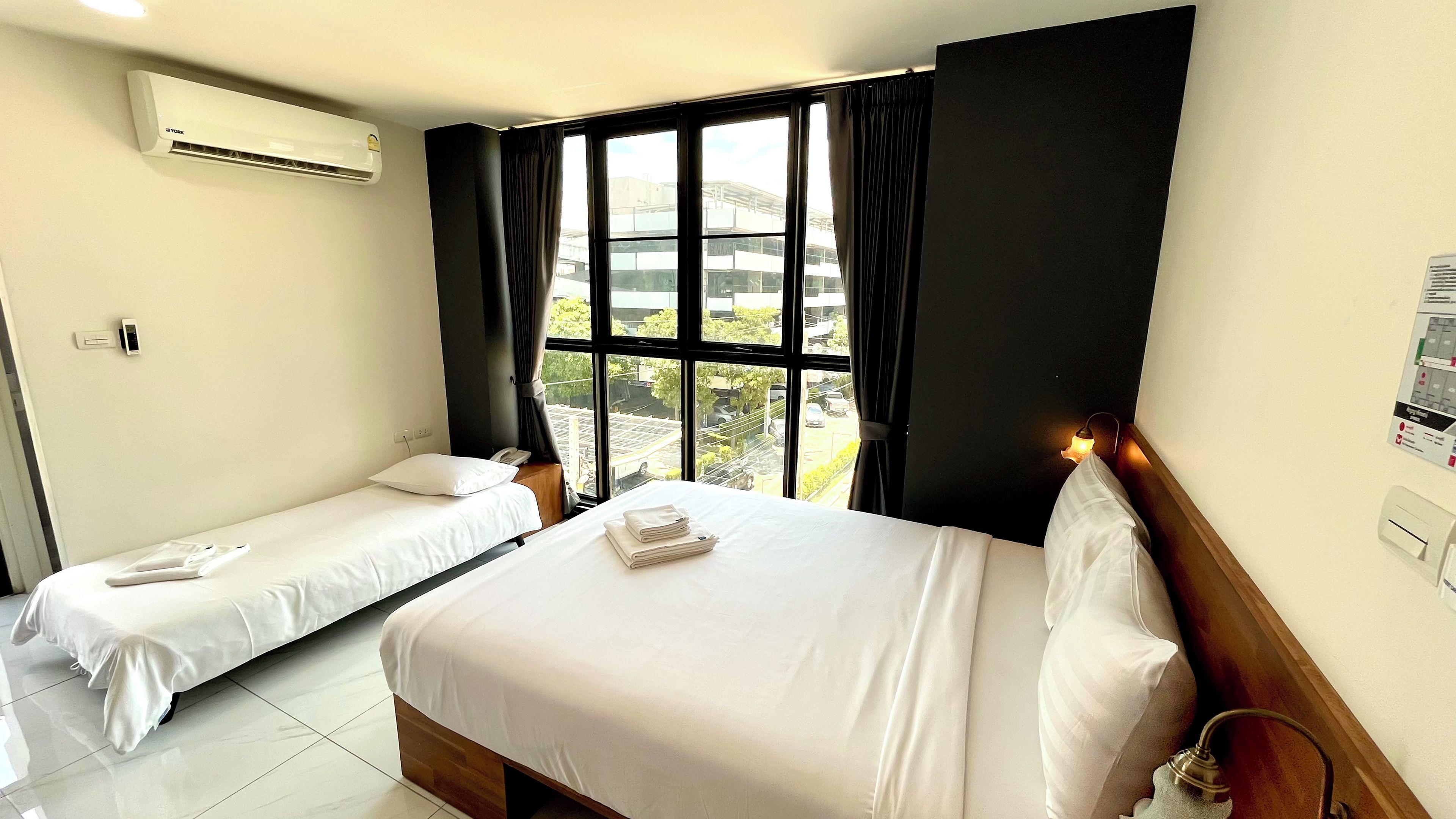 A hotel room with two beds, a window with curtains, an air conditioning unit, and a wall-mounted light fixture.