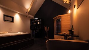 A dimly lit room with a large bathtub, a wooden cabinet, and a small table with a bottle. - Chambre d'hôte with private spa, whirlpool bath and infrared sauna (Boëseghem)