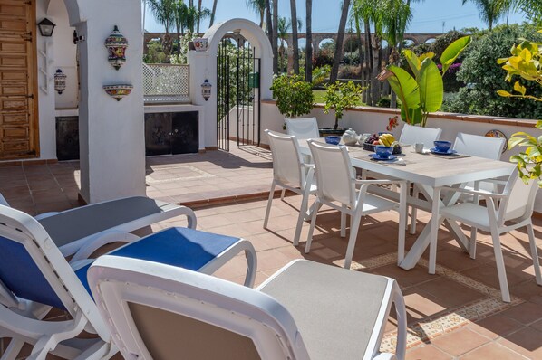 Um pátio externo com móveis brancos, uma mesa de jantar posta com tigelas e xícaras, e vista para o mar.