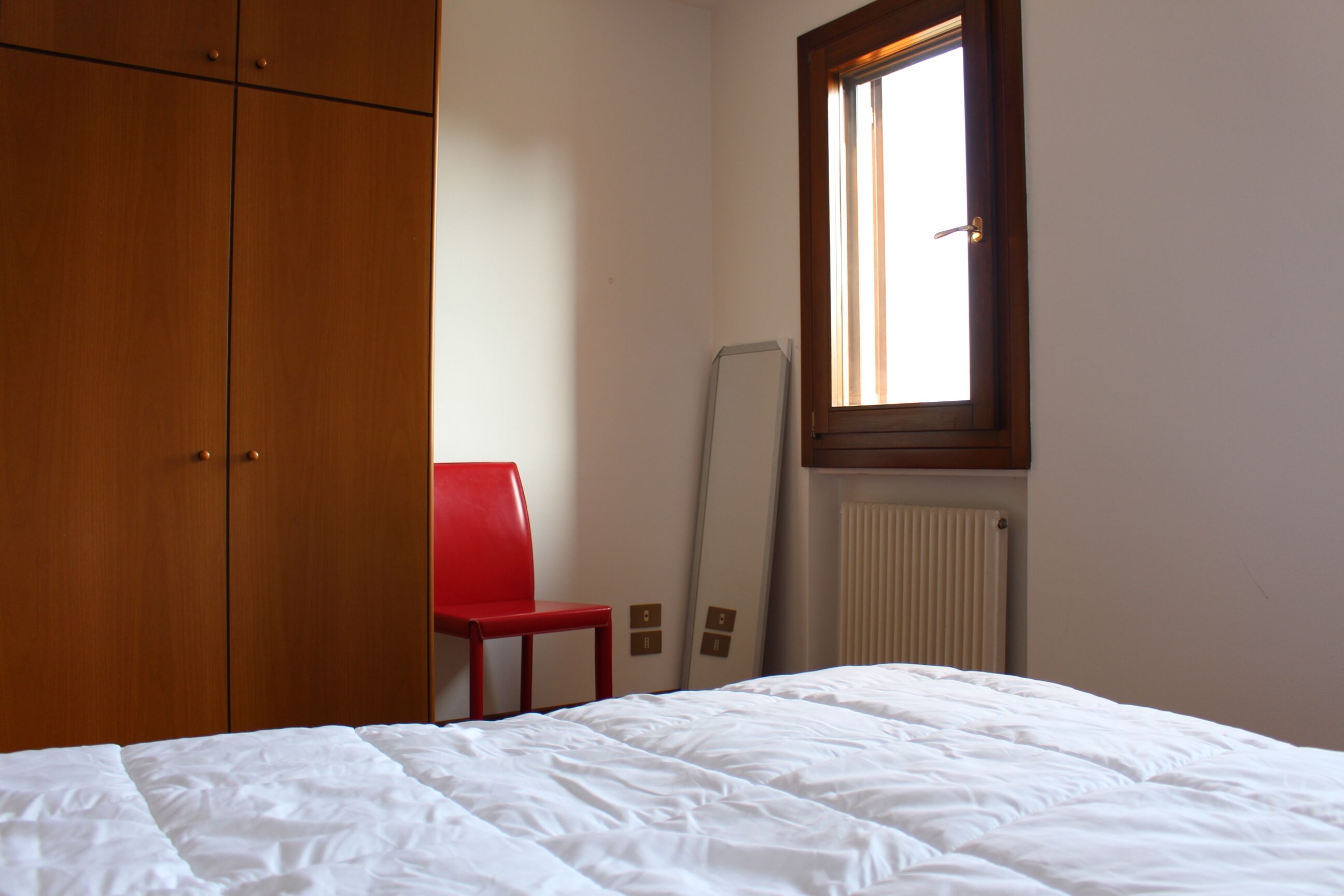 A bedroom with a bed, a red chair, a wardrobe, and a window.