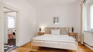 Een slaapkamer met een houten bed, witte wanden, een klein nachtkastje met een lamp, een radiator en een raam met gordijnen.
