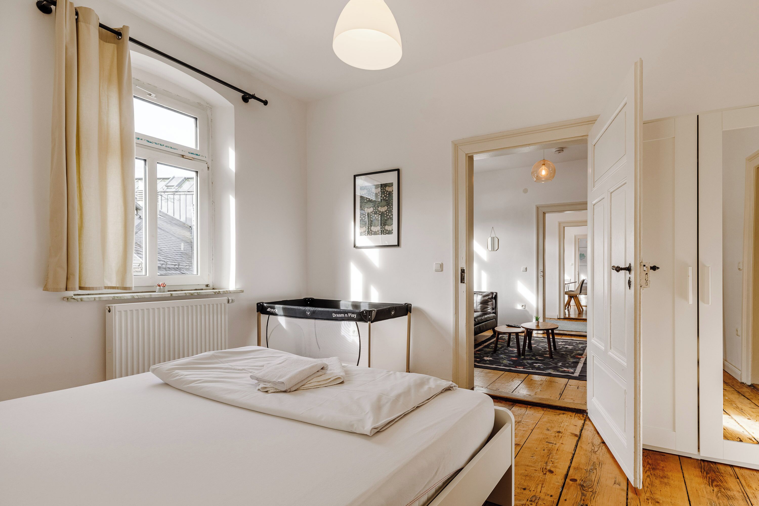 Une chambre à coucher avec un lit, une fenêtre avec des rideaux, un radiateur, un bureau et une porte donnant sur une autre pièce.
