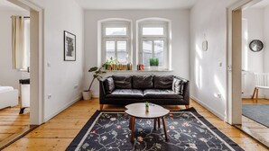 Een woonkamer met een zwarte leren bank, een ronde houten salontafel en een vloerkleed met bloemenprint. Er zijn twee ramen met witte kozijnen, een potplant en een ingelijst kunstwerk aan de muur.
