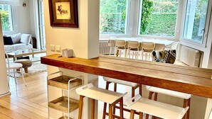 A modern kitchen with a wooden bar counter, white bar stools, and a dining area with white chairs and a table. - Villa de charme à Benerville - 5 Suites, Garden 1800m² & Beach on foot (Benerville-sur-Mer)
