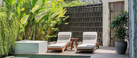 Uma área na beira da piscina com duas espreguiçadeiras, uma mesinha e um vaso de planta.