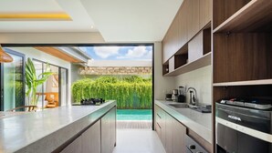 A modern kitchen with a built-in island, a view of a swimming pool, and a brick wall. - Yoki Villa by Betterplace (Canggu)