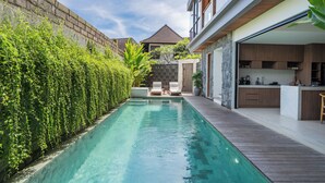 A swimming pool with a wooden deck, a stone wall, and a view of a house with a kitchen and lounge area. - Yoki Villa by Betterplace (Canggu)
