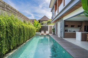 A swimming pool with a wooden deck, a stone wall, and a view of a house with a kitchen and lounge area. - Yoki Villa by Betterplace (Canggu)