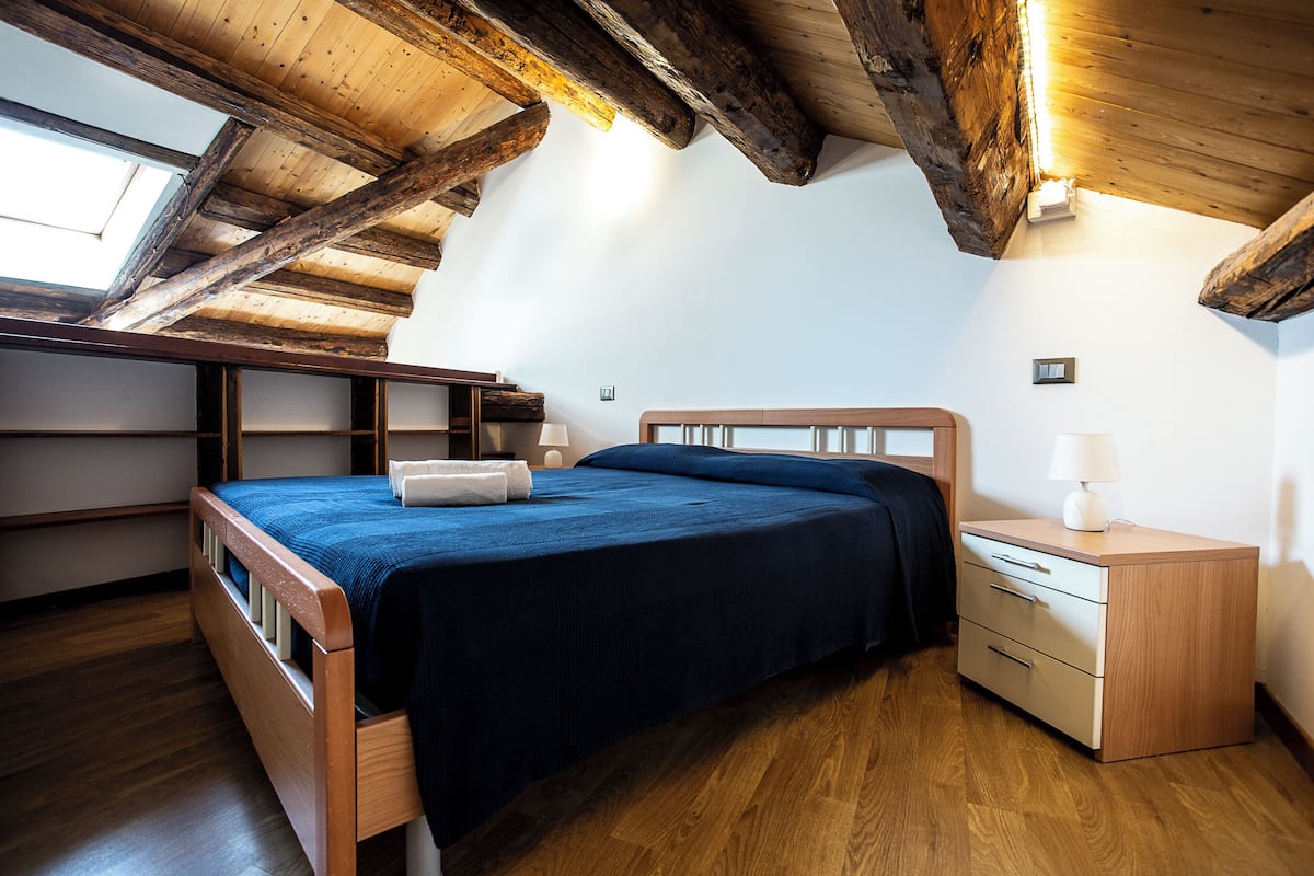 A bedroom with a wooden ceiling, a bed with a blue bedspread, a bedside table with a lamp, and a small window.
