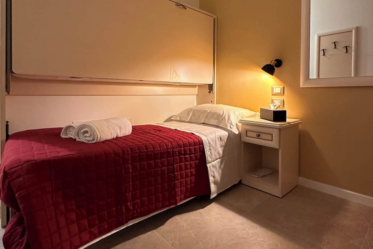 A neatly made bed with a red quilt, a bedside table with a lamp, and a wall-mounted light switch.