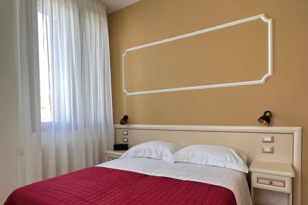 A bedroom with a bed, white curtains, a beige wall, and a nightstand with a lamp.