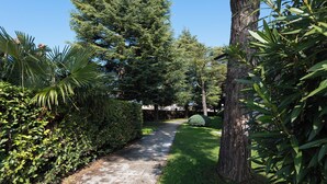 A paved pathway flanked by lush greenery and tall trees, leading towards a building in the distance. - Residence Sant Elena A19 by Wonderful Italy (Peschiera del Garda)