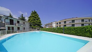 A rectangular swimming pool surrounded by a white fence, with two-story buildings and a large tree in the background. - Residence Sant Elena A19 by Wonderful Italy (Peschiera del Garda)