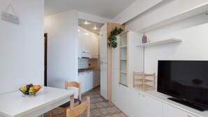 A compact kitchen with white cabinets, a dining table with chairs, a fruit bowl, a television, and a small plant. - Residence Sant Elena A19 by Wonderful Italy (Peschiera del Garda)