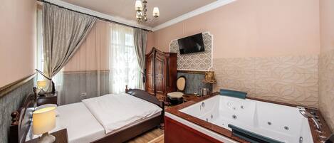 Une chambre avec un grand lit, une table de chevet en bois, une petite table de chevet avec une lampe, une grande baignoire, un téléviseur fixé au mur et un mur décoré d’un motif.