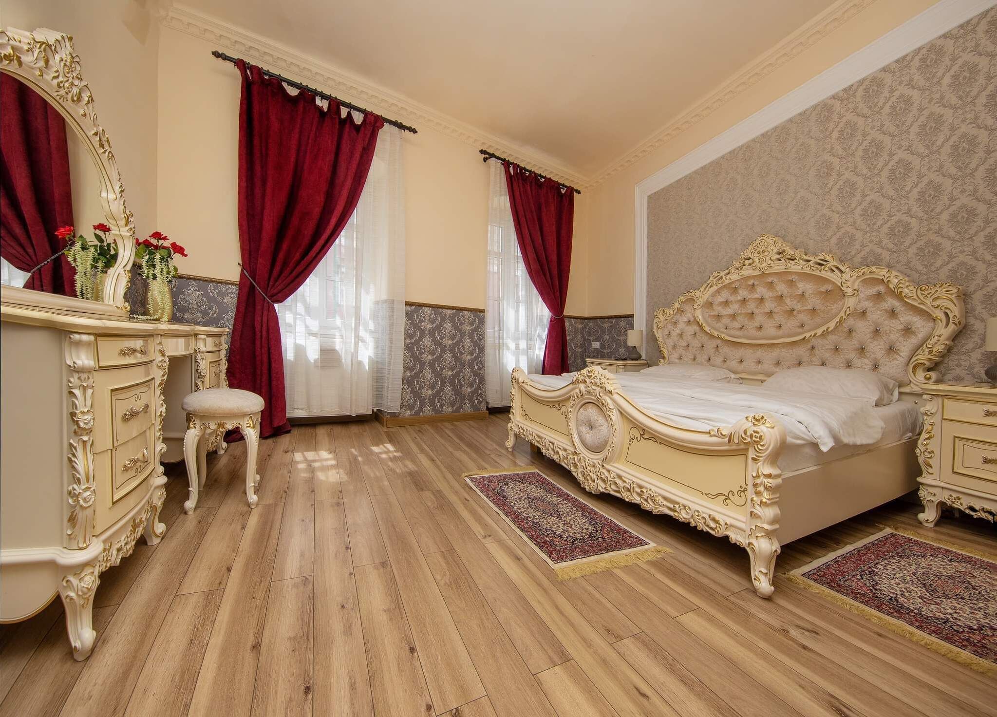 Une chambre à coucher avec un grand lit à baldaquin décoré, une coiffeuse avec un miroir et deux fenêtres ornées de rideaux rouges.