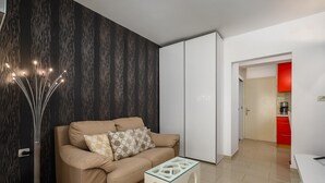 Ein modernes Wohnzimmer mit einem beigen Sofa, einem Glastisch und einer stehenden Stehlampe. Im Hintergrund sind ein Kleiderschrank und ein Küchenbereich zu sehen.