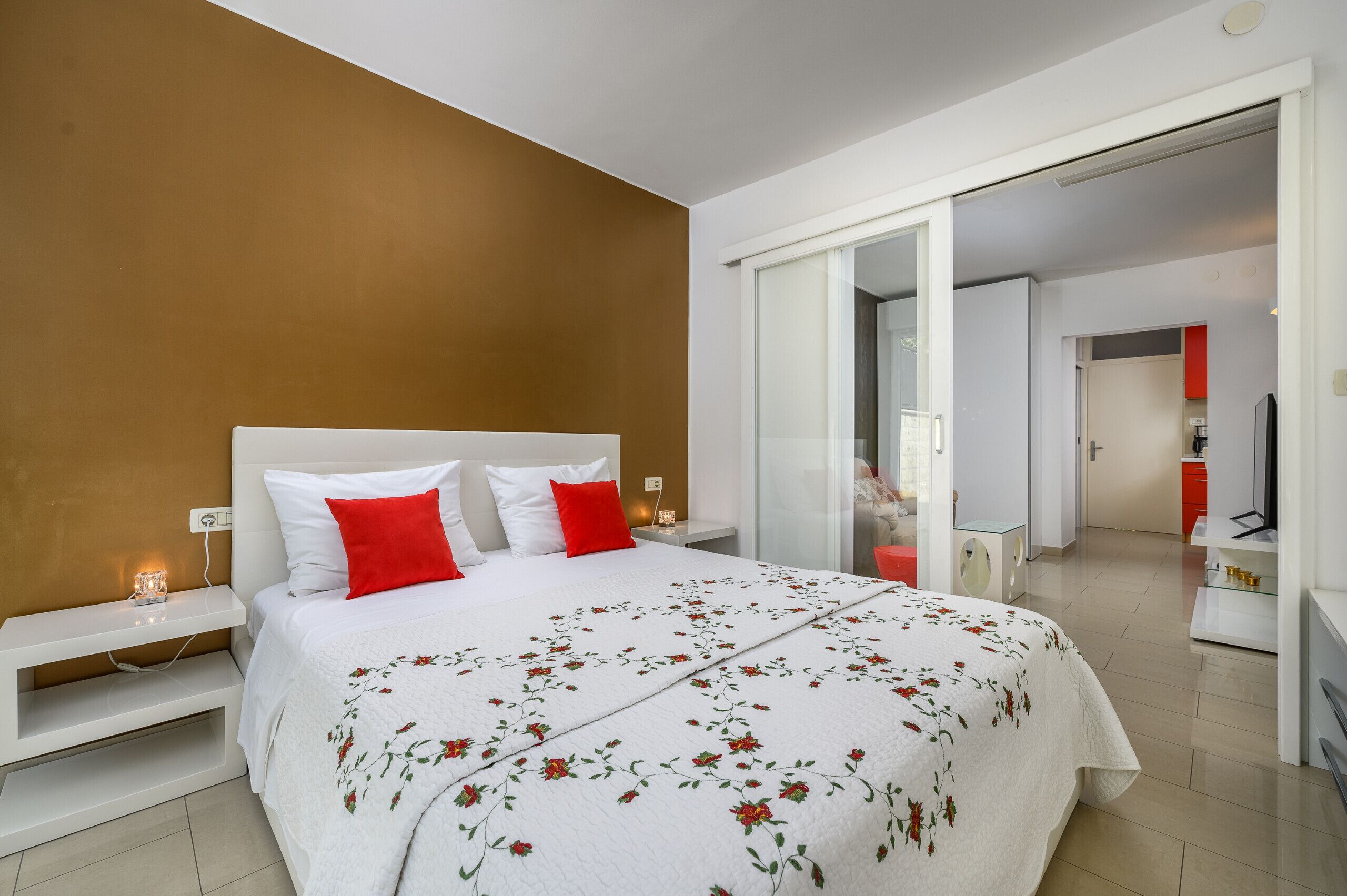 Une chambre d’hôtel moderne avec un grand lit, une table de chevet et un oreiller rouge. Une porte vitrée donne accès à une salle de bain et à un coin cuisine.