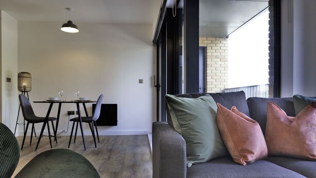 Ein modernes Wohnzimmer mit grauem Sofa, grün-kupferfarbenen Kissen, einem Esstisch mit Stühlen und einem großen Fenster mit Blick auf eine Ziegelwand.