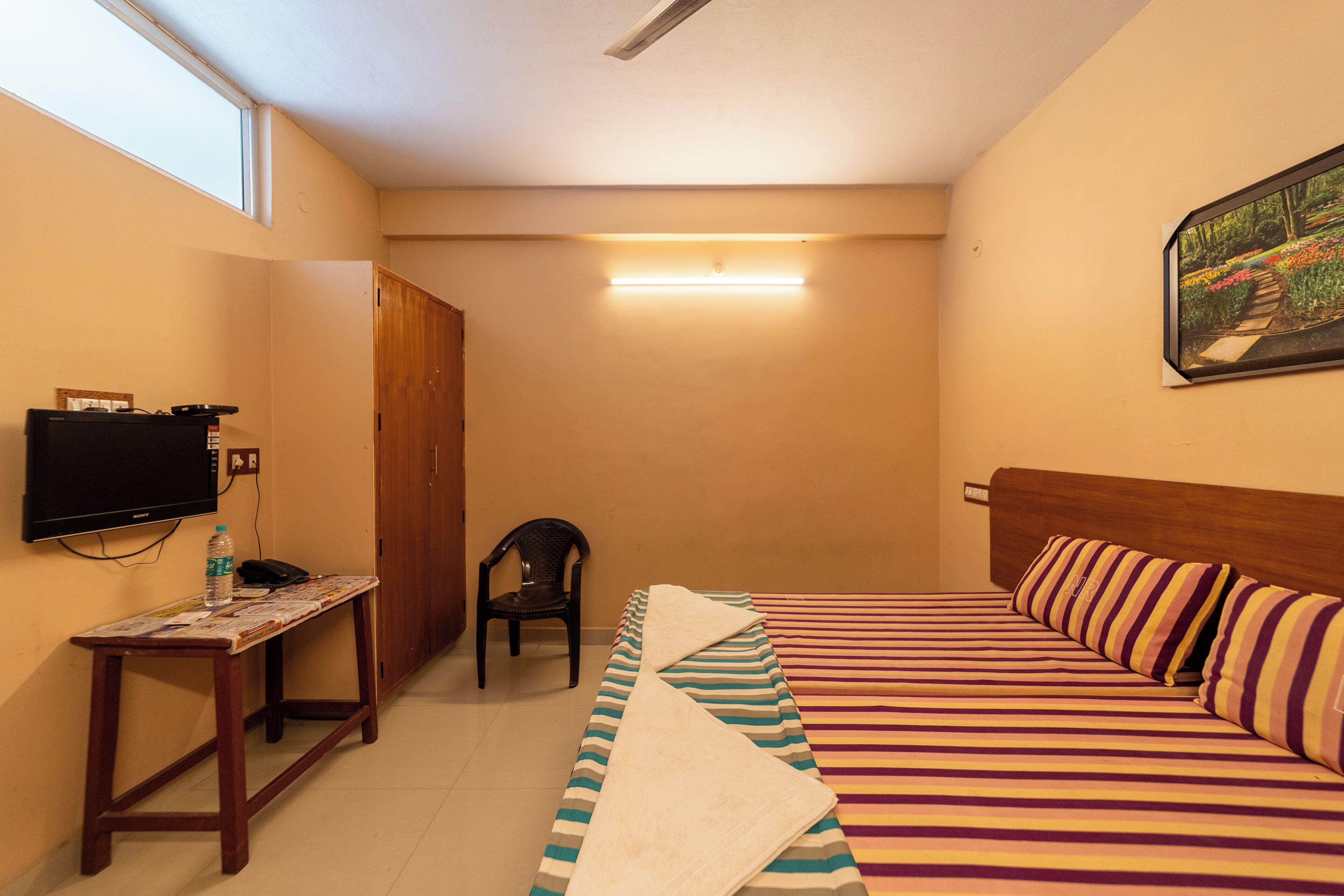 A hotel room with a bed, a TV, a desk with a chair, and a painting on the wall.