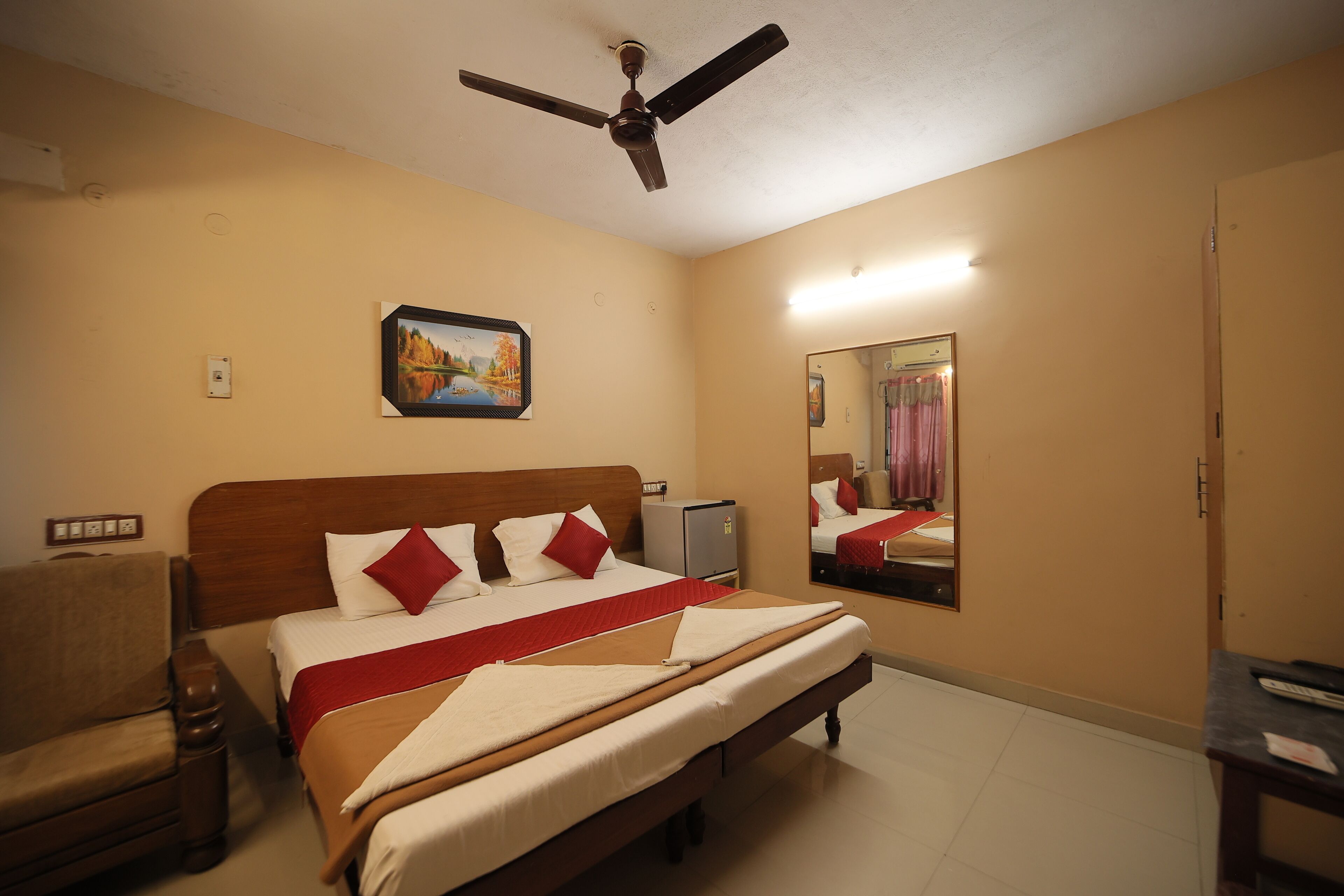 A hotel room with a bed, a chair, a ceiling fan, a painting, a mirror, and a desk.