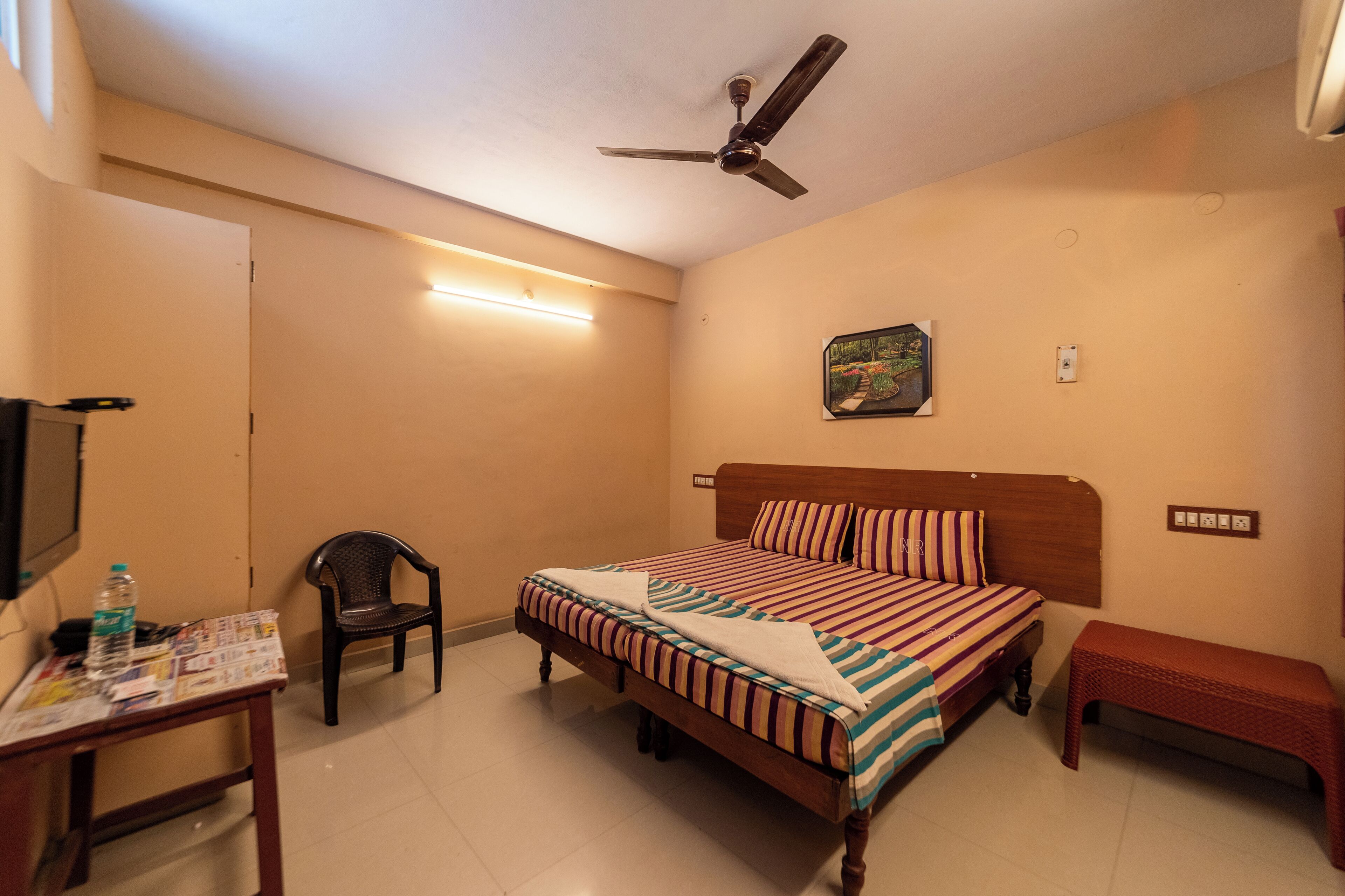 A bedroom with a bed, a chair, a table, a ceiling fan, and a wall-mounted picture.