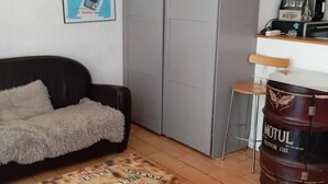 Un espace de vie compact comprenant un canapé, une grande armoire grise, une malle en bois, un tabouret de bar et un bar avec une bouteille d’huile moteur Motul.